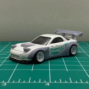K&ouml;nnte beinhalten: Wei&szlig;es Spielzeugauto aus Druckguss mit einem lila Spoiler und dem Aufdruck "Project &mu;" an der Seite. Das Auto steht auf einer gr&uuml;nen Schneidematte.
