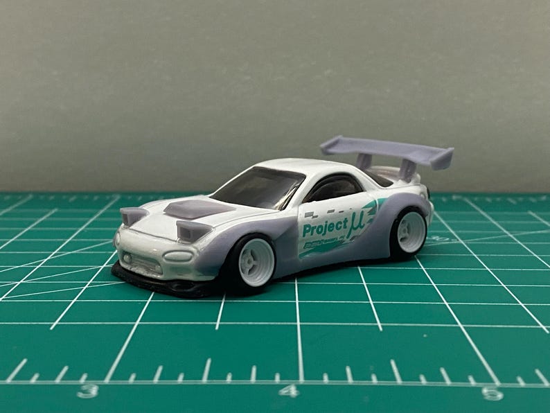 K&ouml;nnte beinhalten: Wei&szlig;es Spielzeugauto aus Druckguss mit einem lila Spoiler und dem Aufdruck "Project &mu;" an der Seite. Das Auto steht auf einer gr&uuml;nen Schneidematte.