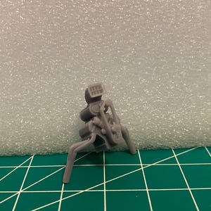 Peut inclure: Un petit modèle gris imprimé en 3D d'un moteur, avec des détails complexes. Le modèle a une section supérieure rectangulaire et plusieurs composants incurvés, avec deux pieds s'étendant vers le bas. Il est posé sur un fond vert quadrillé et blanc.
