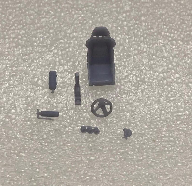 May include: A set of grey plastic model car parts, including a racing seat, steering wheel, exhaust pipes, and a fire extinguisher.