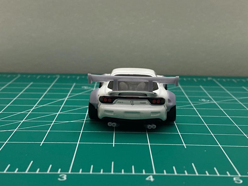 K&ouml;nnte beinhalten: Wei&szlig;es Diecast-Modellfahrzeug mit einem gro&szlig;en Heckfl&uuml;gel. Das Auto hat schwarze R&auml;der und rote R&uuml;ckleuchten. Das Auto steht auf einer gr&uuml;nen Schneidematte.