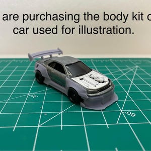 Peut inclure: Une voiture miniature blanche et grise avec un moteur visible. La voiture est sur un tapis de coupe vert. Le texte "You are purchasing the body kit only car used for illustration." est au-dessus de la voiture.