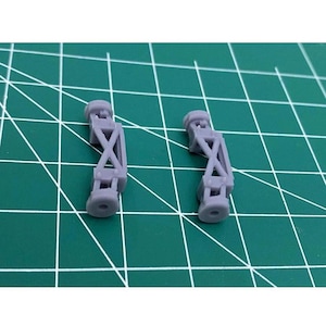 May include: Two small, grey, 3D-printed components. Each has a cylindrical end, an X-shaped support, and a second cylindrical end. The parts are on a green cutting mat with white grid lines.