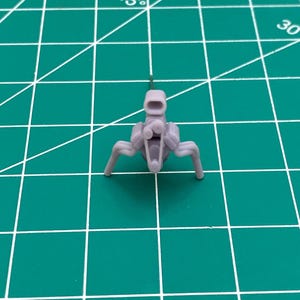 Op de afbeelding: Een grijs plastic model van een kleine motor met vier poten. De motor heeft een kleine uitlaatpijp bovenop.