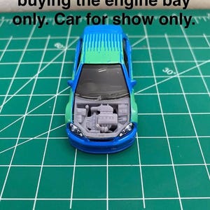 Puede incluir: Un coche de juguete azul y verde con el capó abierto, revelando un compartimento del motor detallado. El texto "Car not included you are buying the engine bay only. Car for show only." es visible sobre el coche.