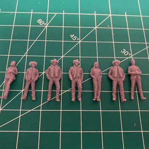 May include: A collection of seven miniature figurines, each depicting a person in various poses. The figures are all the same color and appear to be wearing casual clothing, including jackets, hats, and jeans. The figurines are arranged in a row.