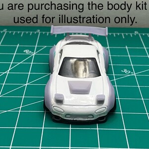 K&ouml;nnte beinhalten: Wei&szlig;es Spielzeugauto aus Druckguss mit einem Bodykit, einem Heckfl&uuml;gel und einem grauen Motorhauben-Scoop. Das Auto befindet sich auf einer gr&uuml;nen Schneidematte.