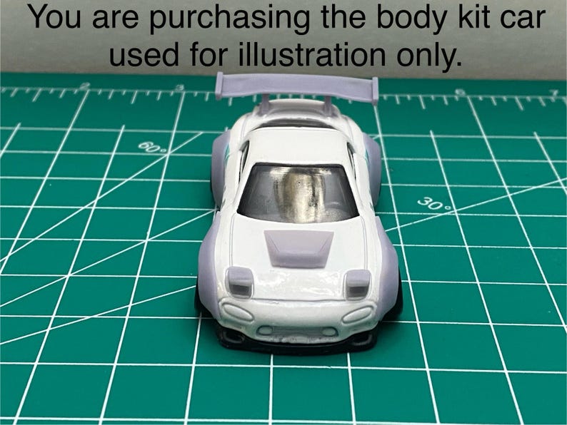 K&ouml;nnte beinhalten: Wei&szlig;es Spielzeugauto aus Druckguss mit einem Bodykit, einem Heckfl&uuml;gel und einem grauen Motorhauben-Scoop. Das Auto befindet sich auf einer gr&uuml;nen Schneidematte.