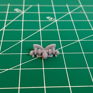 May include: A small, grey, 3D-printed model of an engine component, possibly for a miniature vehicle or model. The component has a complex design with multiple cylinders and tubes. It sits on a green cutting mat.