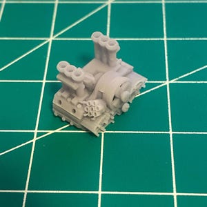 Könnte beinhalten: Ein kleines, graues Modell eines Motors mit mehreren Auspuffrohren und komplizierten Details. Das Modell befindet sich vor einem grünen Gitterhintergrund, was auf eine Werkstatt oder Bastelumgebung hindeutet. Das Motormodell ist wahrscheinlich für ein Miniaturfahrzeug oder ein Ausstellungsstück.