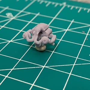 May include: A small, light gray model of an engine, featuring intricate details of the cylinders, exhaust manifolds, and other components. The engine model is mounted on a small, beige base. The background is a green cutting mat with white grid lines.