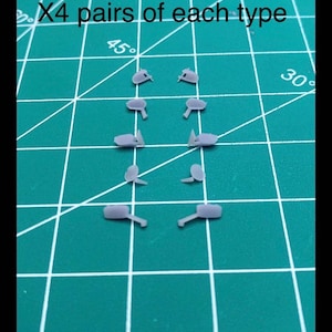 May include: A collection of small, gray plastic model parts arranged on a green grid surface. The text "X4 pairs of each type" is visible. The parts appear to be components for a model kit or similar project.