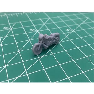 May include: A small, gray motorcycle model with detailed features, including a front headlight, handlebars, and a visible engine. The miniature bike is positioned on a green cutting mat with grid lines, suggesting a crafting or hobby context.