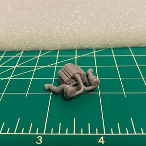 May include: A small, gray model of an engine, featuring intricate details like tubes and cylinders. The engine is positioned on a green cutting mat with white grid lines. The background includes a white foam block.