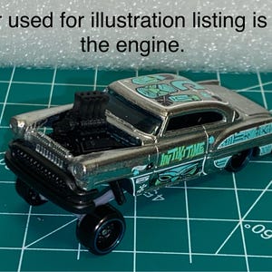 Op de afbeelding: Een zilveren speelgoedauto met een zwarte motor en zwarte wielen. De auto heeft turquoise en witte graphics op het dak en de zijkanten, met de tekst "In Tiki Time". De tekst "Car used for illustration listing is for the engine" staat boven de auto.