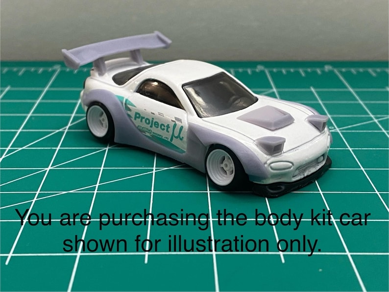 K&ouml;nnte beinhalten: Wei&szlig;es Diecast-Modellauto mit einem grauen Bodykit, einem Heckfl&uuml;gel und dem Schriftzug "Project &mu;" an der Seite. Das Auto steht auf einer gr&uuml;nen Schneidematte.