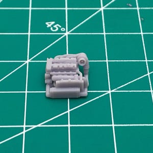 May include: A small, gray, plastic model of a car engine. The engine has a detailed design with multiple cylinders and a pipe coming out of the top.