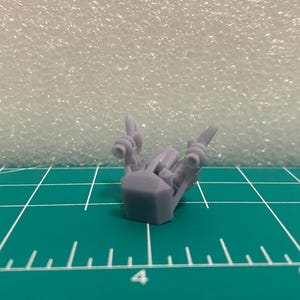 Peut inclure: Une petite figurine miniature en résine imprimée en 3D de couleur violet clair, dotée d'une base hexagonale centrale et de deux bras s'étendant vers le haut, chacun se terminant par une ouverture circulaire sur un tapis de découpe vert.