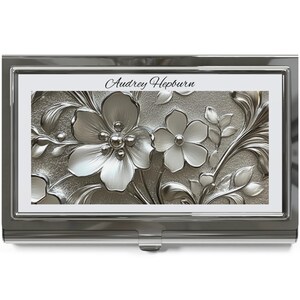 Pode incluir: Um porta-cartões prateado com um design floral. O design apresenta flores e folhas em relevo com um acabamento metálico. O texto "Audrey Hepburn" está impresso no topo.