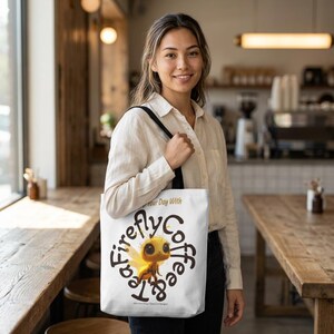 Könnte beinhalten: Weiße Tragetasche mit schwarzen Trägern. Sie zeigt eine Cartoon-Glühwürmchen-Illustration und die Worte "Tea Firefly Coffee" in einem kreisförmigen Design. Das Glühwürmchen ist gelb und braun mit großen Augen. Der Text "Start Your Day With" steht darüber.