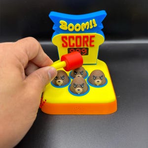 May include: A colorful Whac-A-Mole game with a red and yellow mallet. The game features four brown mole characters, a digital score display, and the words "BOOM!! SCORE". The game is yellow, blue, and orange.
