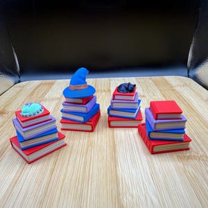 Puede incluir: Varias pilas de libros en miniatura, coloridos, con un sombrero de bruja azul y un gato negro. Los libros tienen páginas rojas, azules y moradas, con cubiertas rojas y azules. El sombrero tiene una banda marrón.