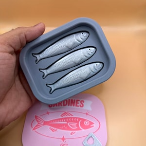 May include: A gray sardine-shaped tray with three silver fish inside, held above a pink sardine can lid. The lid features a pink sardine illustration and the word "SARDINES" in a banner.