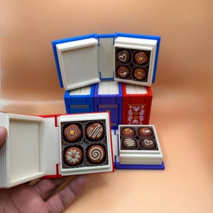 Op de afbeelding: Miniatuur boekvormige dozen in blauw, rood en paars, elk met decoratieve chocolade. De chocolade is bruin met witte en gele details. De dozen zijn ontworpen om eruit te zien als open boeken, met de chocolade erin.