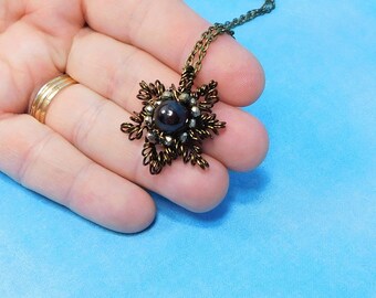 May include: A bronze-toned wire-wrapped pendant necklace featuring a central dark round bead surrounded by intricate metallic loops and small dark accents. The pendant is attached to a matching bronze-colored metal chain link necklace.