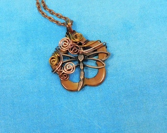Puede incluir: Un collar con colgante de libélula de color cobre. El colgante presenta un diseño de libélula con detalles de alambre y acentos en espiral en tonos dorados y cobrizos. El collar tiene una cadena delicada y está sobre un fondo azul.