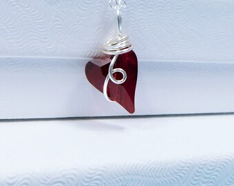 Puede incluir: Un collar de plata con un colgante en forma de corazón de color rojo intenso. El colgante está envuelto en alambre de plata. El collar se muestra sobre cajas blancas texturizadas.