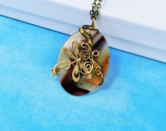Puede incluir: Un collar con un colgante de ágata envuelto en alambre dorado con un diseño floral. El colgante tiene forma ovalada y tiene colores marrón, blanco y dorado.