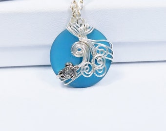 May include: A silver necklace with a blue glass pendant featuring a wire-wrapped sea turtle design. The pendant is surrounded by silver wire swirls, creating a wave-like effect.