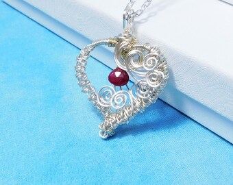 May include: A silver-toned wire-wrapped heart pendant with a small, round, red gemstone. The pendant hangs from a delicate silver chain. The heart design features intricate swirls and is displayed against a white box on a blue background.