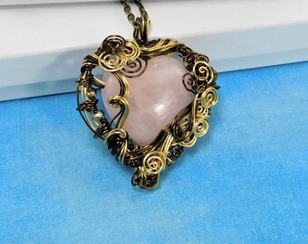 May include: A heart-shaped rose quartz pendant encased in intricate, dark bronze-toned wire wrapping. The wire features multiple decorative swirls and coils that frame the smooth, pale pink stone. A matching dark metal chain is attached at the top.
