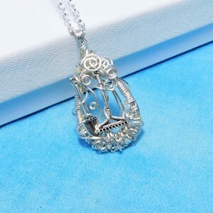 May include: A silver necklace with a pendant shaped like a teardrop. The pendant is made of wire and features a design of gardening tools, including a shovel, rake, and trowel.