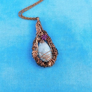Puede incluir: Un collar con un colgante en forma de l&aacute;grima, con una piedra blanca envuelta en alambre de cobre. El colgante tiene un peque&ntilde;o adorno de flor morada.