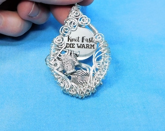 May include: A silver-toned wire-wrapped pendant necklace featuring a circular metal charm engraved with the text Knit Fast DIE WARM. The pendant includes a small three-dimensional charm of a ball of yarn and knitting needles, all suspended on a thin silver-toned chain.