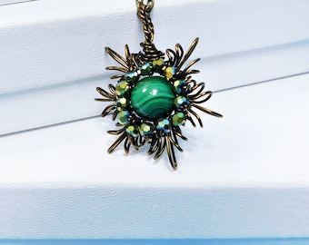 May include: A pendant necklace featuring a round, green, banded malachite stone at the center, surrounded by a ring of small, iridescent green beads and an intricate, star-shaped frame of twisted bronze-toned wire on a matching chain.