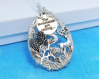 May include: A silver-toned wire-wrapped pendant necklace featuring a circular charm engraved with the text The mountains are calling... along with metal accents shaped like a deer, pinecones, and foliage, all suspended from a delicate silver chain.