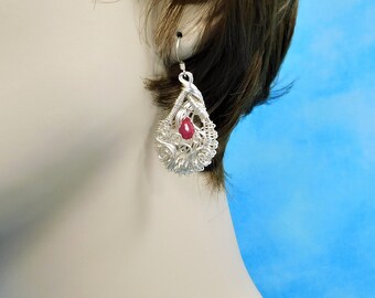 May include: A silver filigree teardrop earring with a red gemstone center. The earring is suspended from a silver hook. The filigree design is intricate and detailed, with a delicate, openwork pattern.