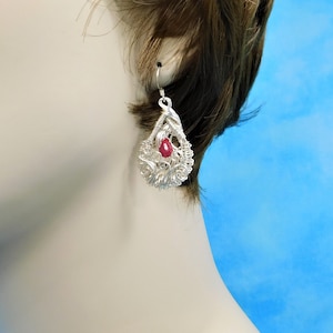 May include: A silver filigree teardrop earring with a red gemstone center. The earring is suspended from a silver hook. The filigree design is intricate and detailed, with a delicate, openwork pattern.