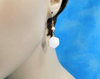 May include: A drop earring featuring a small white spherical bead connected to a larger white hexagonal pendant. The earring is suspended from a thin silver-toned metal hook, showcasing a clean design with a smooth, polished finish on the white components.