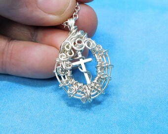 May include: A silver-colored wire-wrapped pendant featuring a central cross motif encased within a circular frame of intricate, swirling metal wire loops. The pendant is attached to a delicate silver-colored chain link necklace.