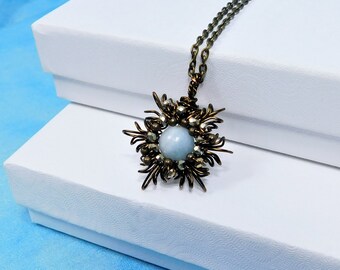 May include: A necklace featuring a central round light blue stone surrounded by dark metallic wire work and small dark beads. The pendant is attached to a dark bronze-toned chain, creating a star-like or floral shape with intricate wire loops.