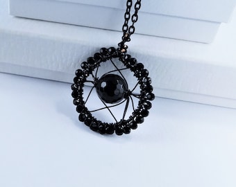 May include: A black necklace featuring a circular pendant constructed from wire and small black beads. A single, larger, faceted black spherical bead is suspended in the center of the circle by thin wire spokes, attached to a matching black chain.