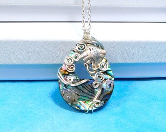 May include: A teardrop-shaped abalone shell pendant featuring an iridescent surface with swirling green, blue, and purple hues. The shell is encased in silver-toned wire wrapping with a small metal stingray figure attached to the top section.
