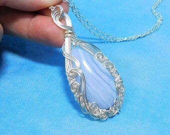 May include: A teardrop-shaped blue lace agate pendant wrapped in intricate silver wire, suspended from a delicate silver chain. The stone features soft, light blue and white banding, secured by a decorative wire-wrapped metal frame.
