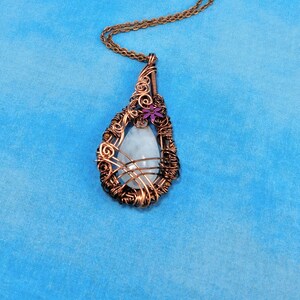 Puede incluir: Un collar con un colgante de alambre de cobre envuelto alrededor de una piedra preciosa blanca con un acento de flor morada.