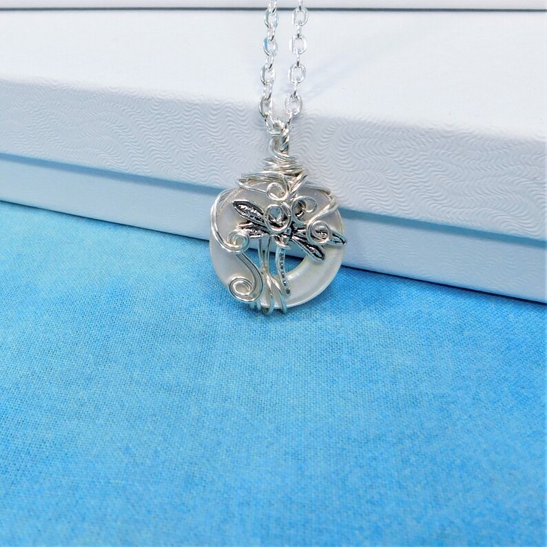 May include: A silver necklace with a white iridescent pendant featuring a dragonfly design. The pendant is made of wire wrapped around a round piece of glass.